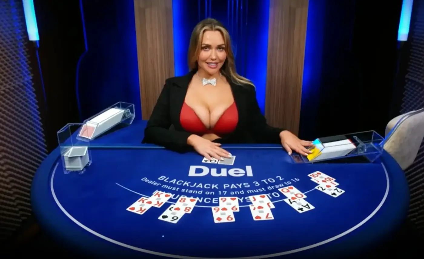 Mia Malkova at the Duel Blackjack Live table in a black blazer, smiling behind the blue felt with Duel branding while cards are dealt mid-stream